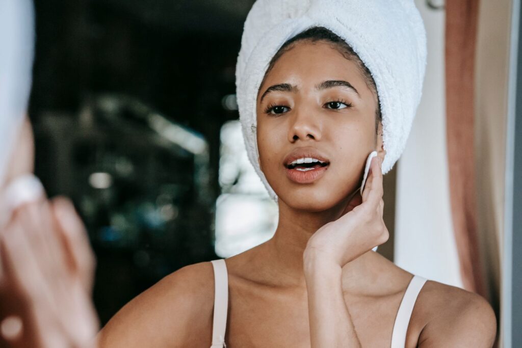 Mulher com toalha na cabeça aplicando produto facial com algodão